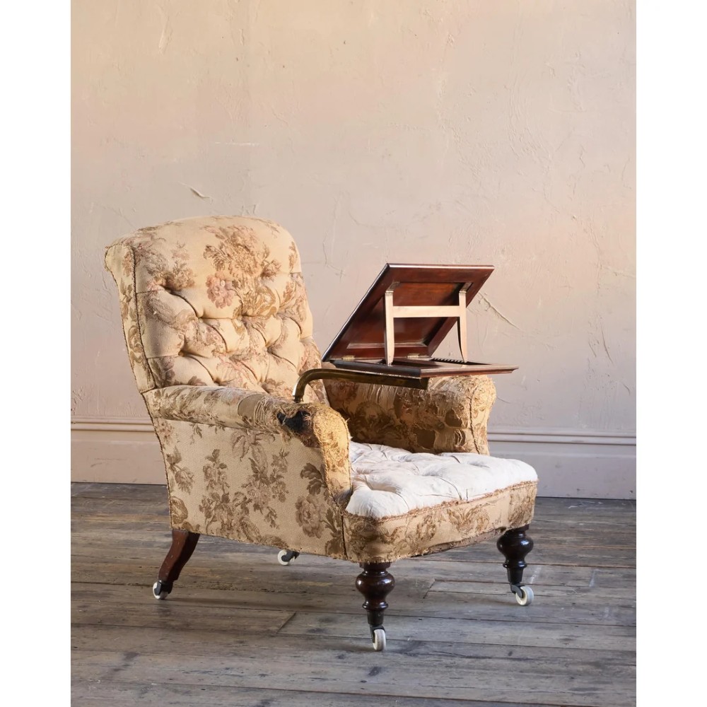 A 19th-century Victorian library armchair upholstered in beige floral pattern fabric with deep buttoning, turned front legs on castors, and an attached adjustable mahogany reading stand.