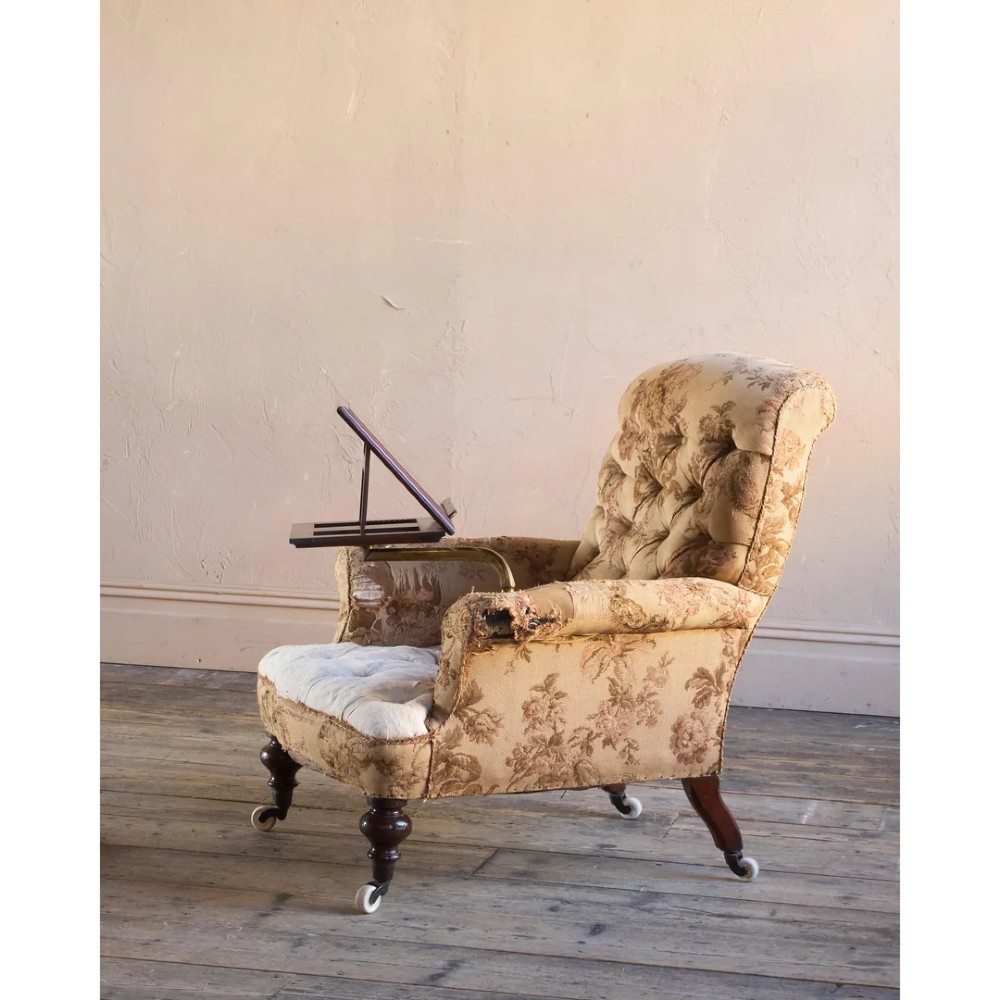 A 19th-century Victorian library armchair upholstered in beige floral pattern fabric with deep buttoning, turned front legs on castors, and an attached adjustable mahogany reading stand. View II