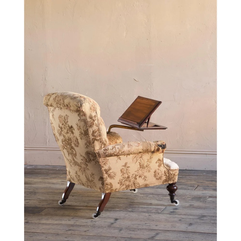 A 19th-century Victorian library armchair upholstered in beige floral pattern fabric with deep buttoning, turned front legs on castors, and an attached adjustable mahogany reading stand. View VII