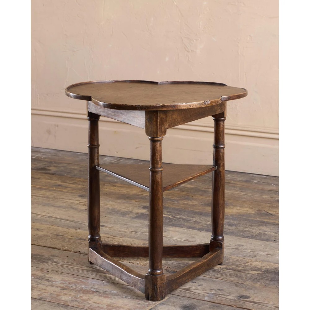 An 18th-century dark oak cricket table featuring a trefoil-shaped top, three turned legs, a triangular lower shelf, and a stretcher base.