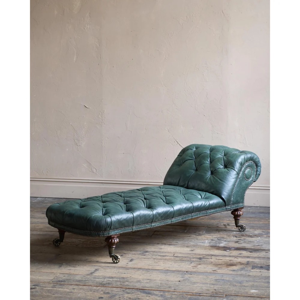 A deep buttoned green leather chaise longue featuring brass stud details, turned wood legs, and brass castors.