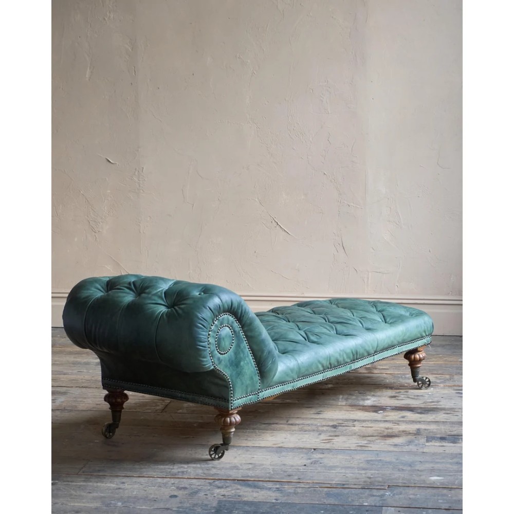 A deep buttoned green leather chaise longue featuring brass stud details, turned wood legs, and brass castors. View III