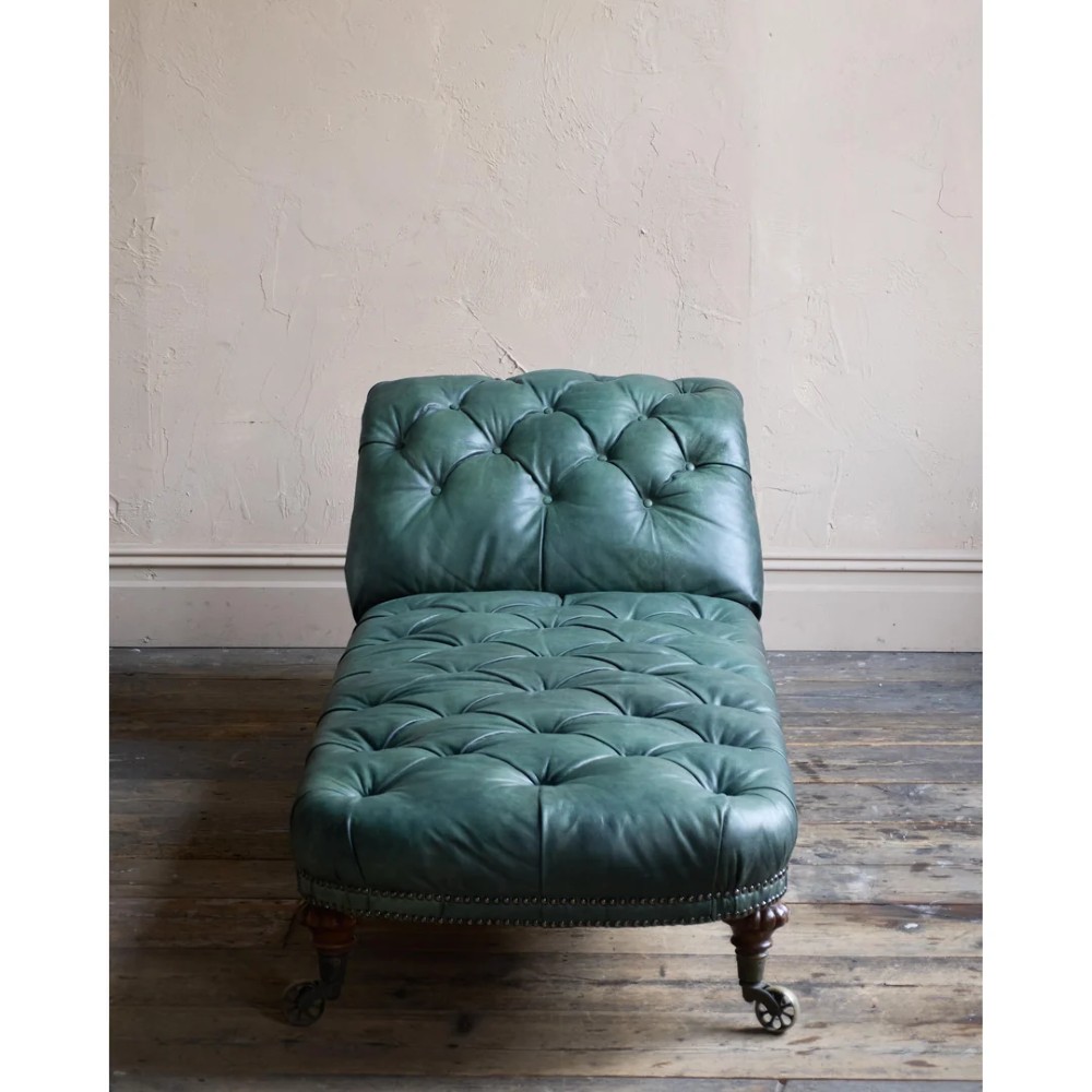 A deep buttoned green leather chaise longue featuring brass stud details, turned wood legs, and brass castors. View V