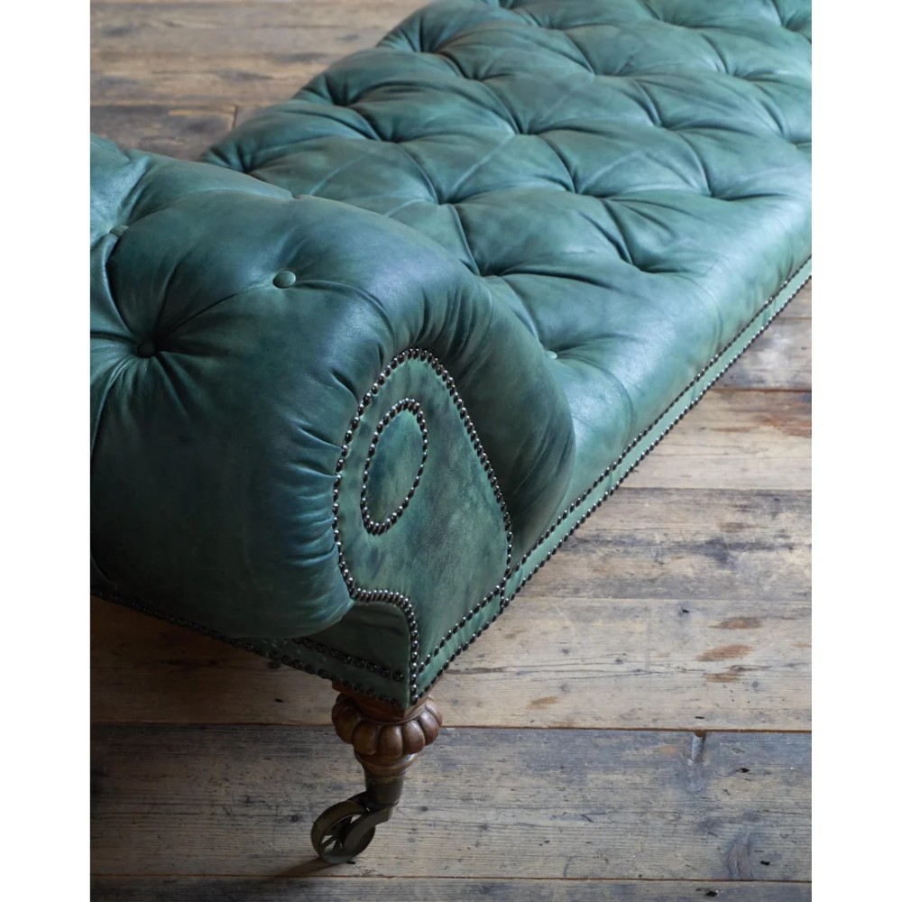 A deep buttoned green leather chaise longue featuring brass stud details, turned wood legs, and brass castors. View VI