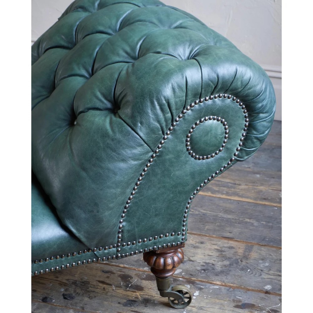 A deep buttoned green leather chaise longue featuring brass stud details, turned wood legs, and brass castors. View VII
