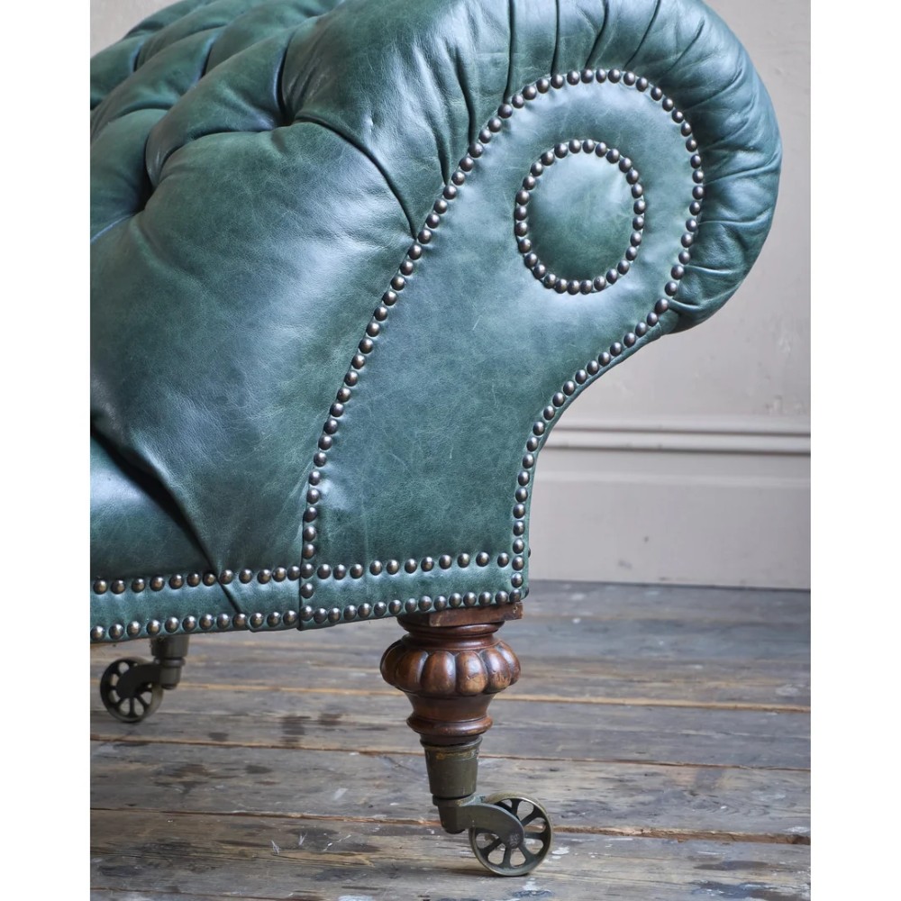 A deep buttoned green leather chaise longue featuring brass stud details, turned wood legs, and brass castors. View X