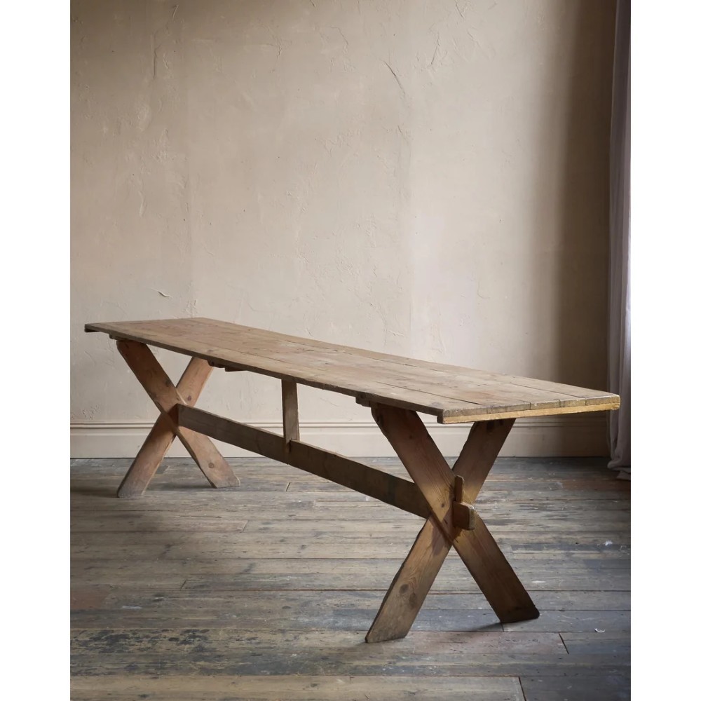 A long, narrow rustic wooden dining table featuring a thick plank top supported by X-frame trestle legs and a longitudinal stretcher.