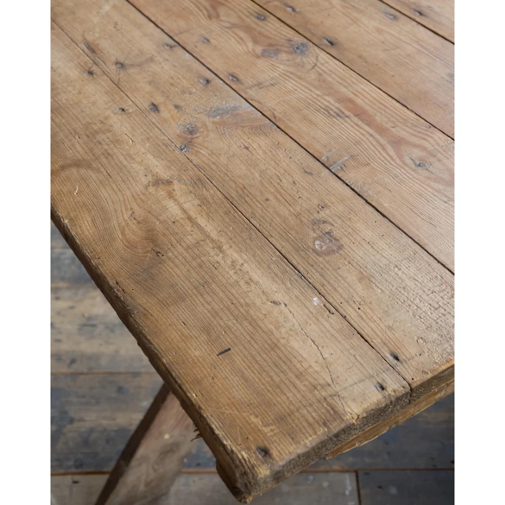 A long, narrow rustic wooden dining table featuring a thick plank top supported by X-frame trestle legs and a longitudinal stretcher. View X