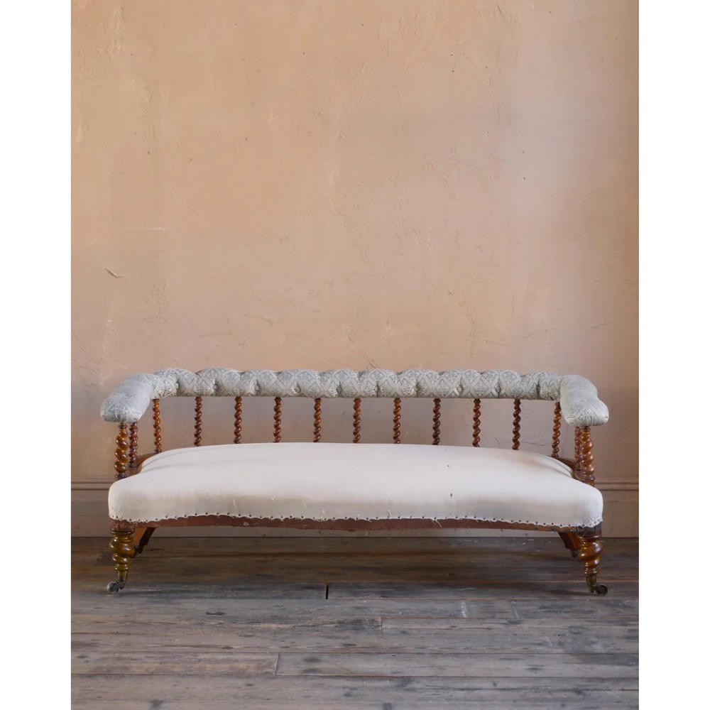 A nineteenth-century salon sofa featuring a curved button-upholstered crest rail supported by turned spindles, a cream padded seat, and turned mahogany legs on brass castors. View V