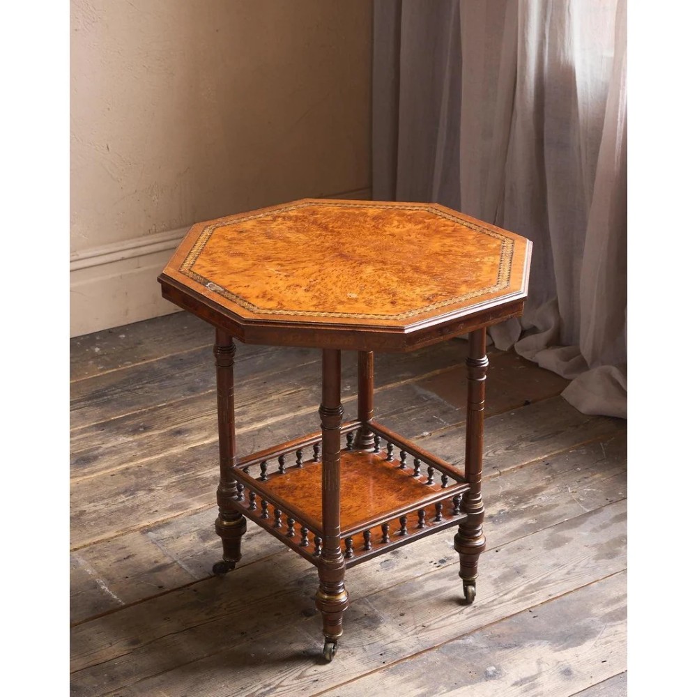 An octagonal Victorian side table featuring an amboyna wood veneer top with gilt borders, turned legs, a lower shelf with a spindle gallery, and brass castors.