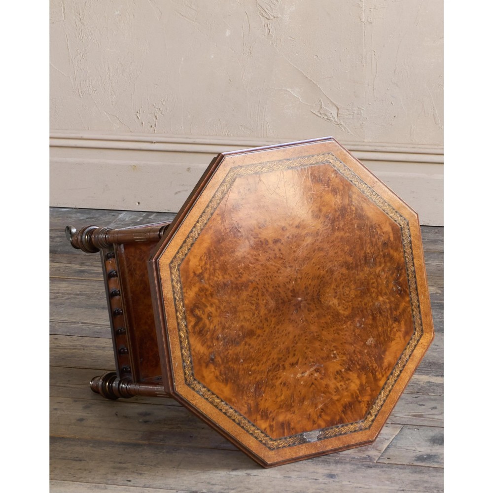 An octagonal Victorian side table featuring an amboyna wood veneer top with gilt borders, turned legs, a lower shelf with a spindle gallery, and brass castors. View IV