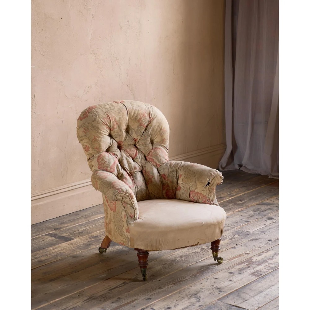 An antique Victorian spoon-back armchair with deep button tufting, worn floral upholstery, turned mahogany front legs, and brass castors.