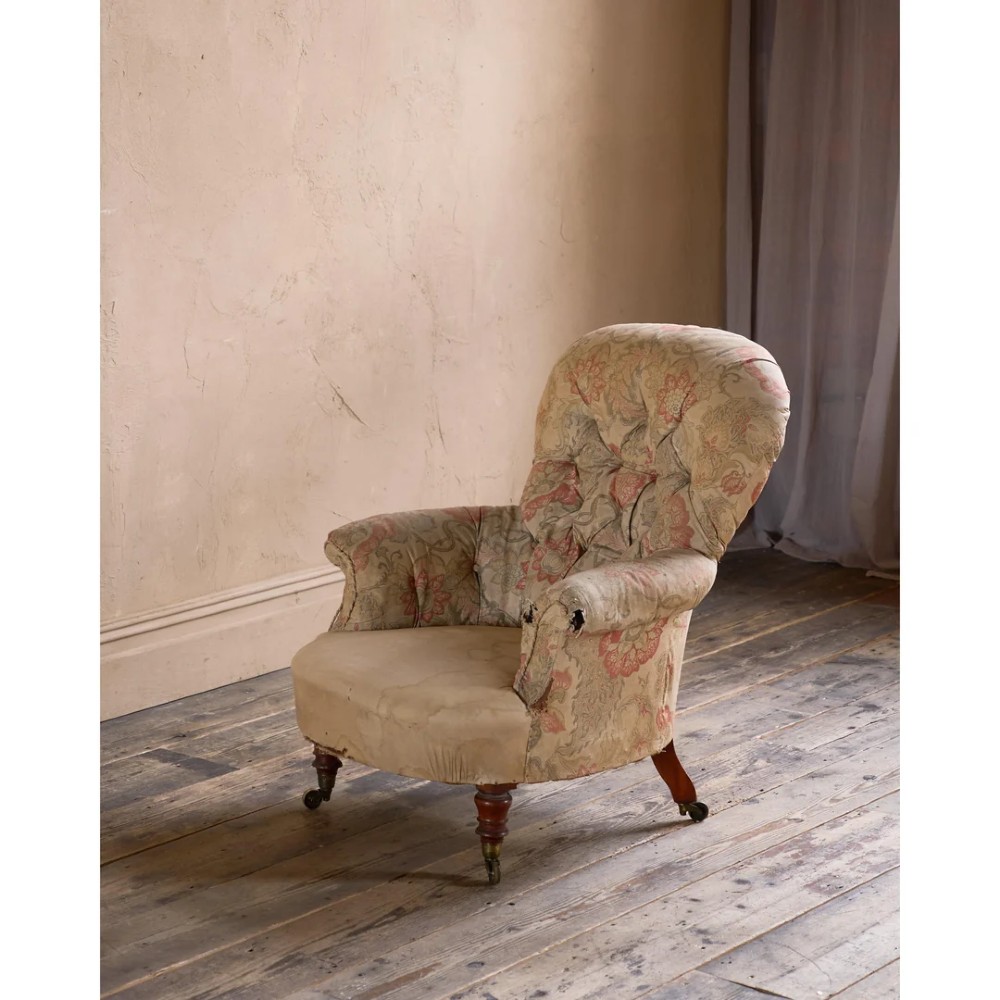 An antique Victorian spoon-back armchair with deep button tufting, worn floral upholstery, turned mahogany front legs, and brass castors. View II