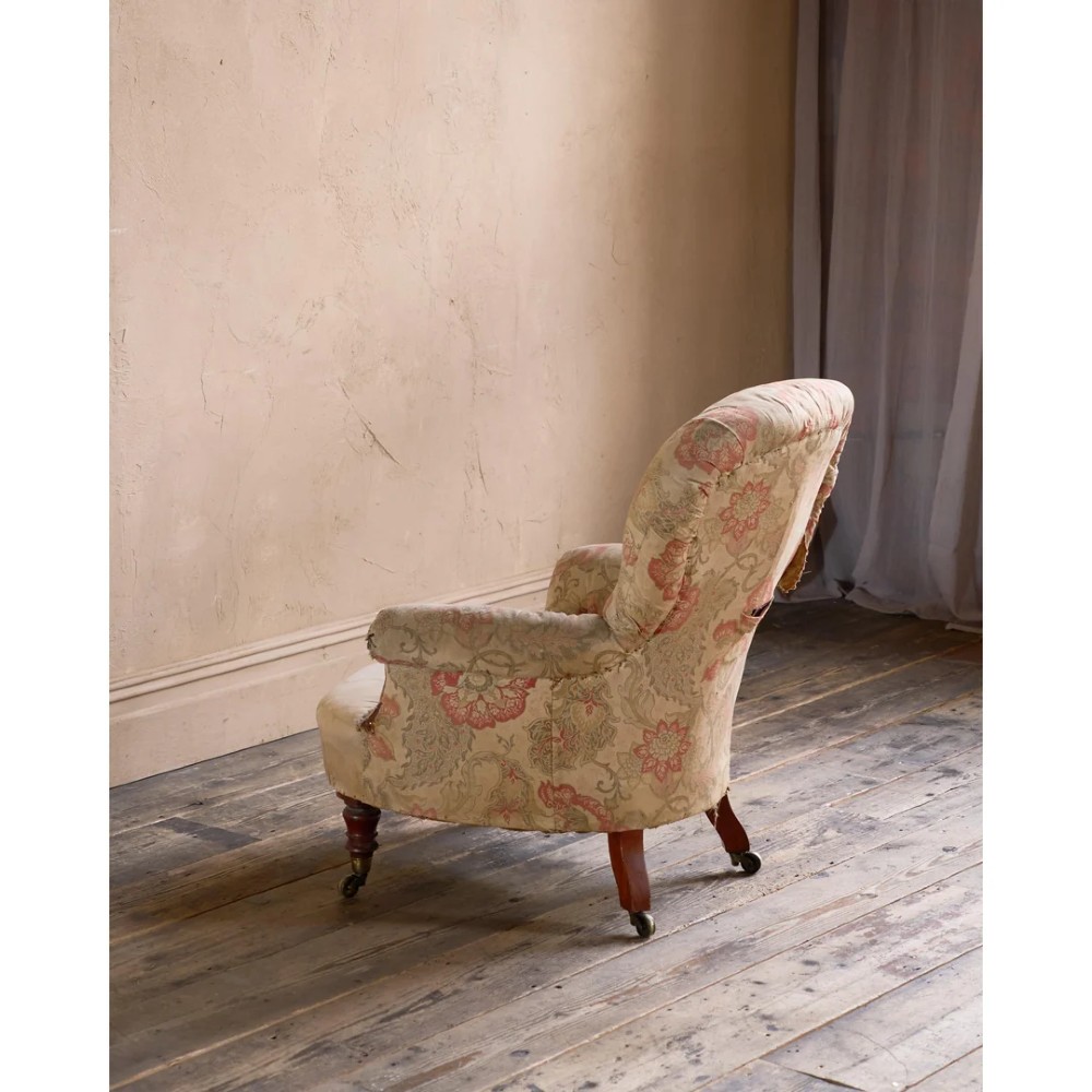 An antique Victorian spoon-back armchair with deep button tufting, worn floral upholstery, turned mahogany front legs, and brass castors. View IV