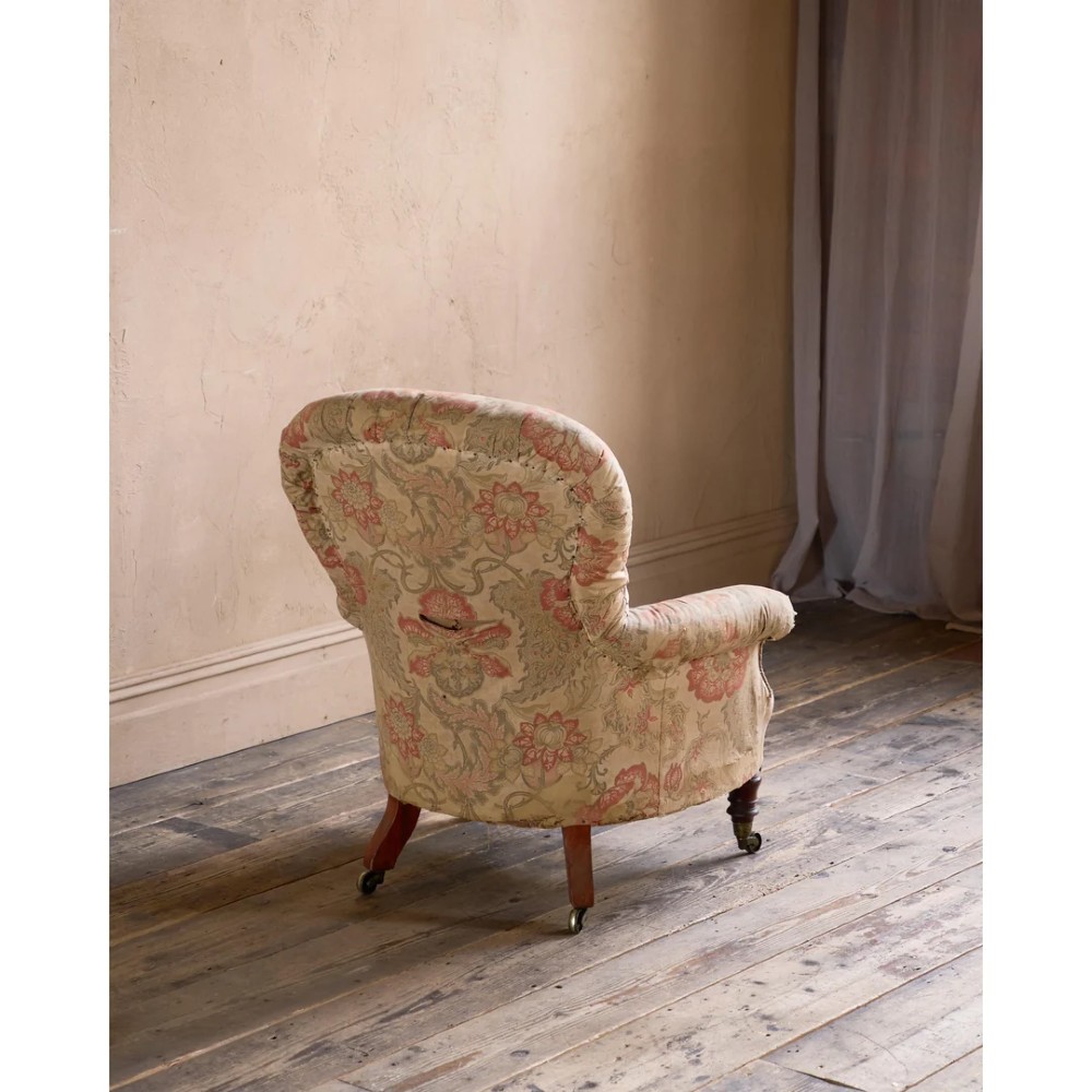 An antique Victorian spoon-back armchair with deep button tufting, worn floral upholstery, turned mahogany front legs, and brass castors. View V