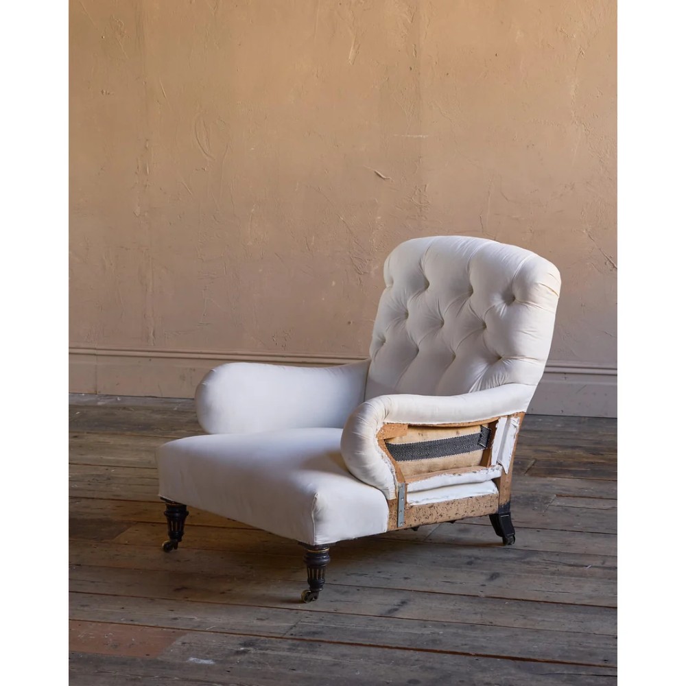 A deep-seated Howard style armchair upholstered in cream calico fabric with a button-tufted backrest and turned wooden legs on brass castors.