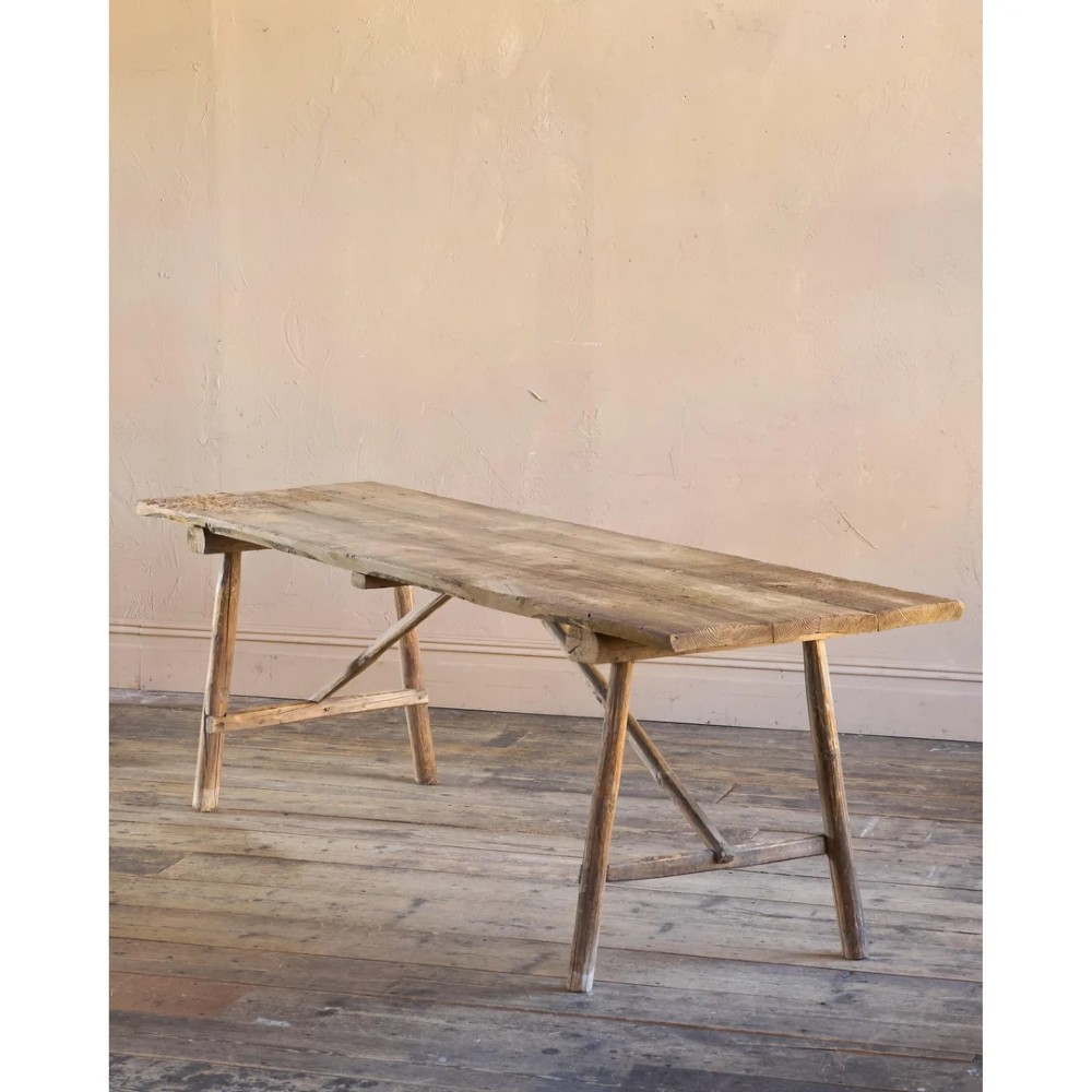 A primitive rustic dining table constructed from weathered pale timber with a rough-hewn plank top and diagonal cross-braced legs.