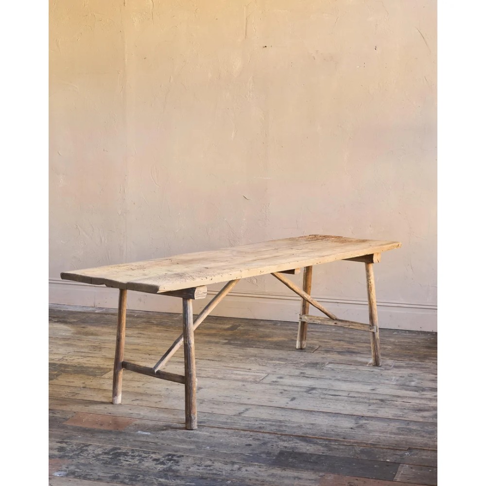 A primitive rustic dining table constructed from weathered pale timber with a rough-hewn plank top and diagonal cross-braced legs. View IV
