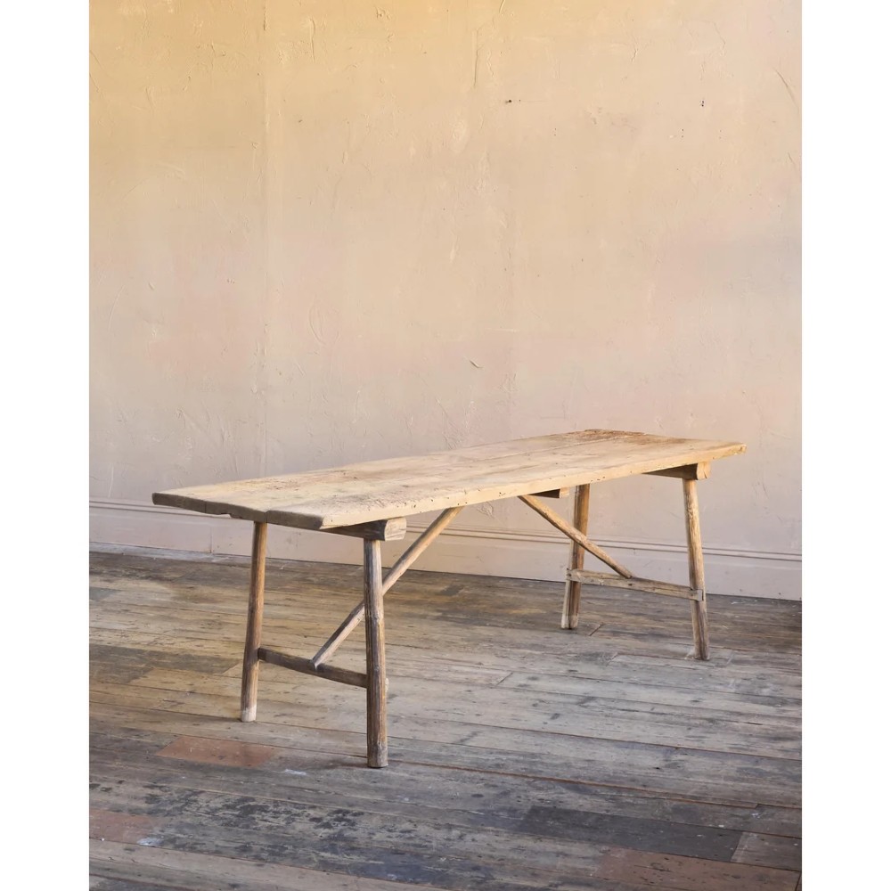 A primitive rustic dining table constructed from weathered pale timber with a rough-hewn plank top and diagonal cross-braced legs. View V