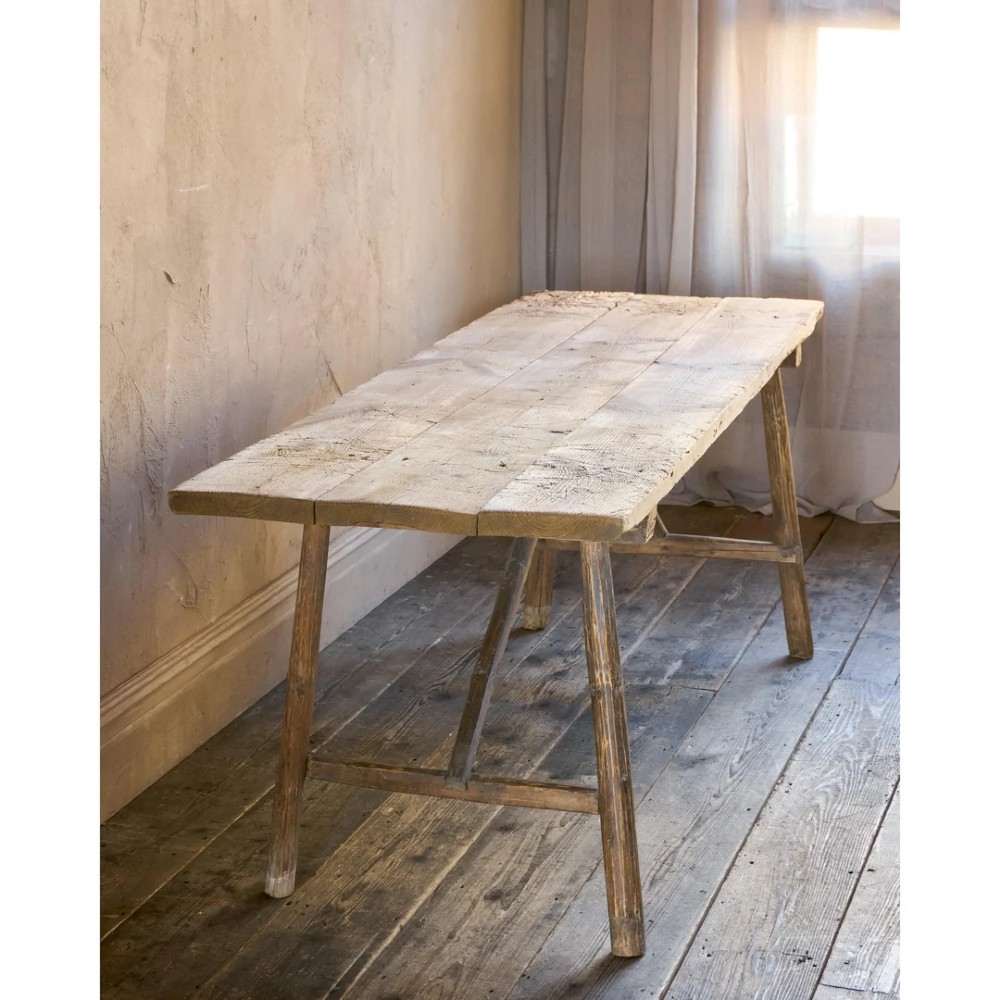 A primitive rustic dining table constructed from weathered pale timber with a rough-hewn plank top and diagonal cross-braced legs. View VII