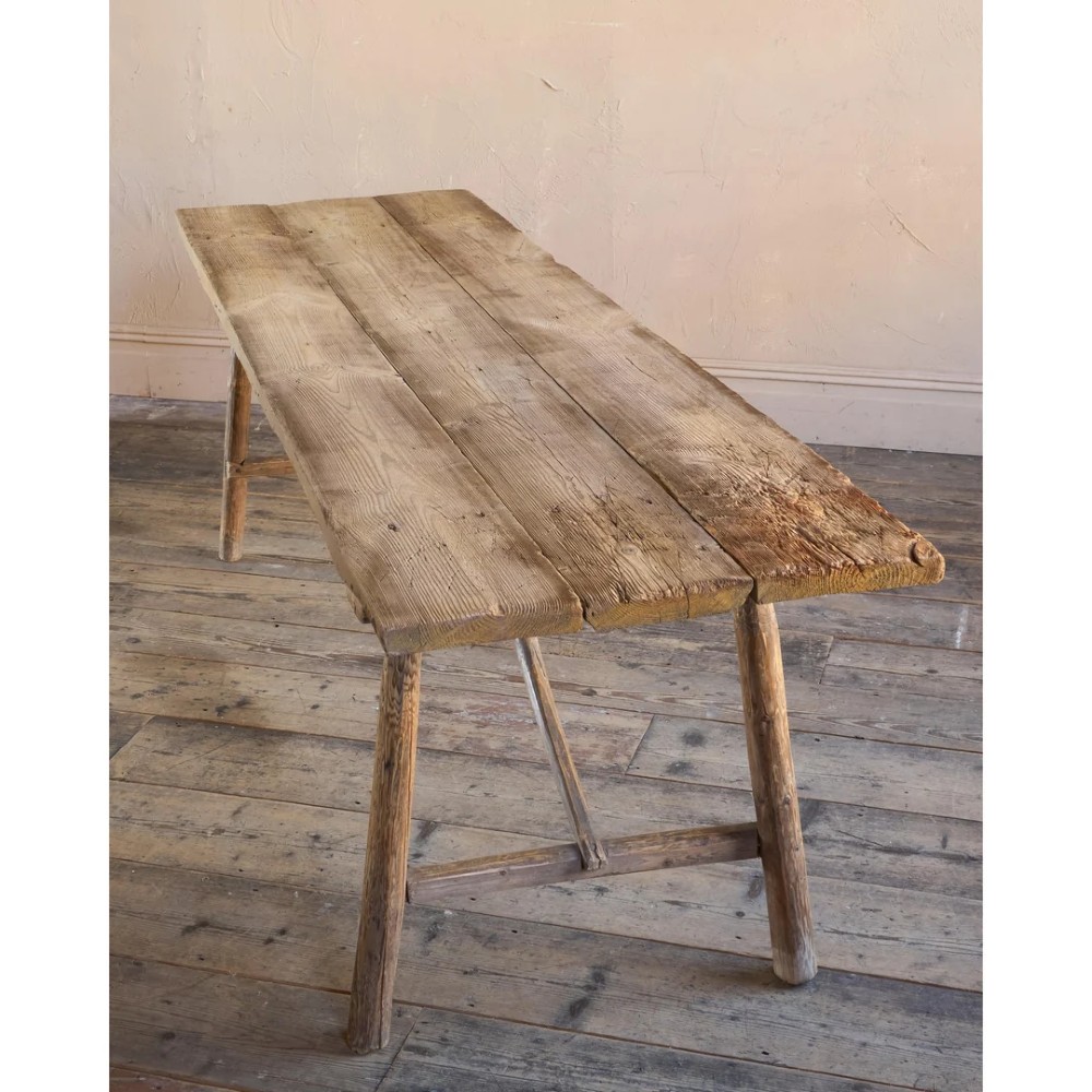 A primitive rustic dining table constructed from weathered pale timber with a rough-hewn plank top and diagonal cross-braced legs. View VIII