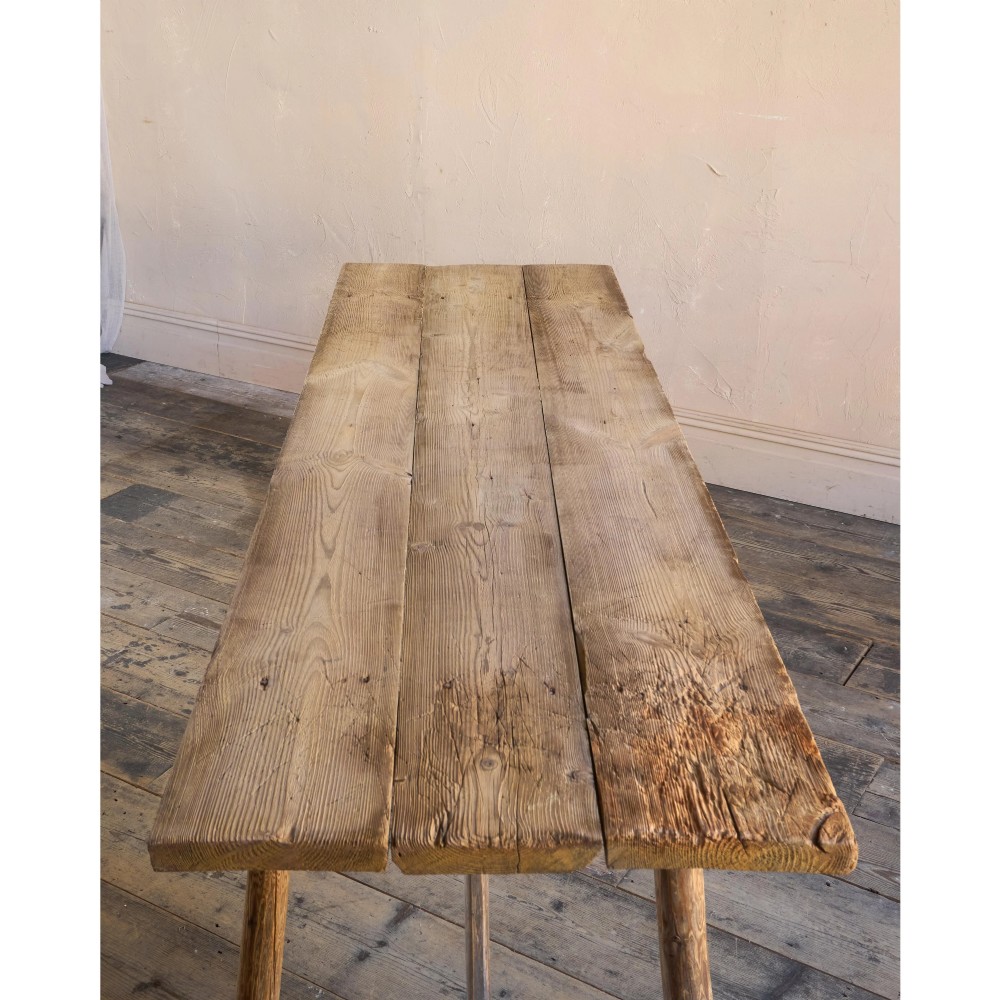 A primitive rustic dining table constructed from weathered pale timber with a rough-hewn plank top and diagonal cross-braced legs. View X
