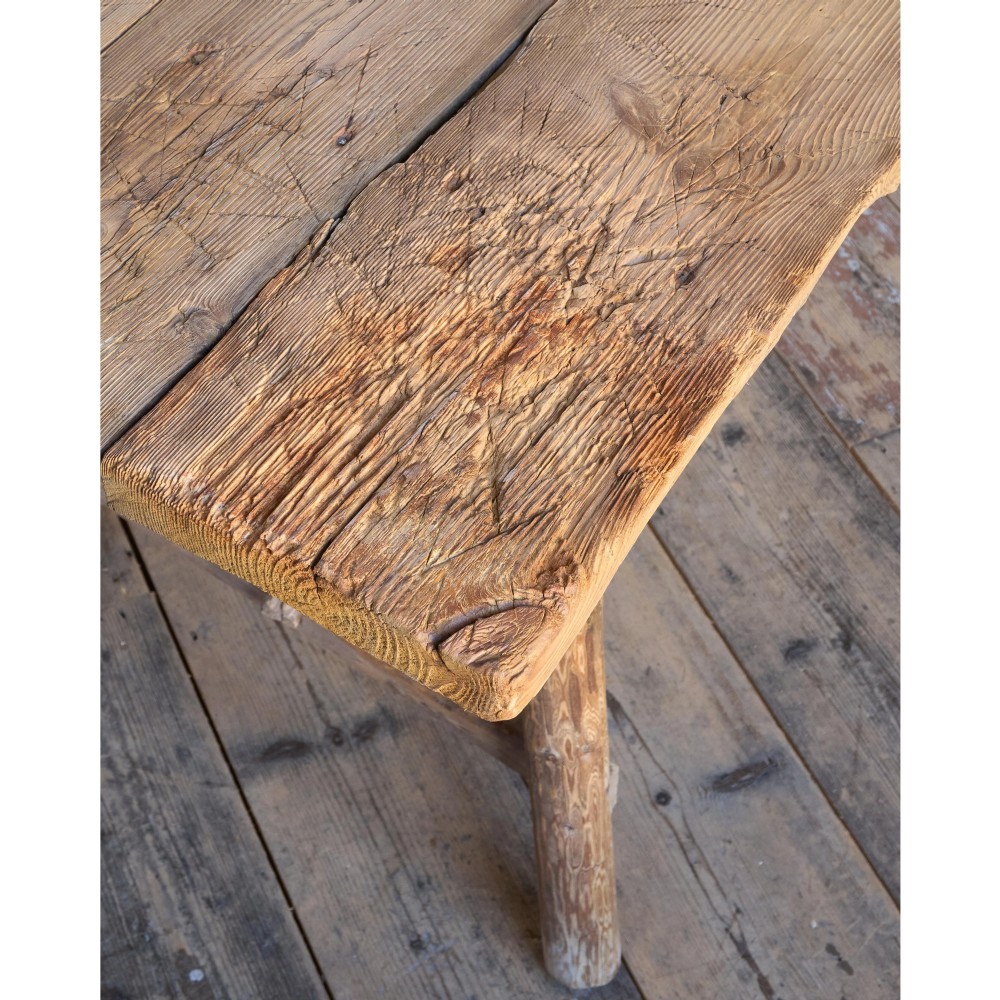 A primitive rustic dining table constructed from weathered pale timber with a rough-hewn plank top and diagonal cross-braced legs. View XII