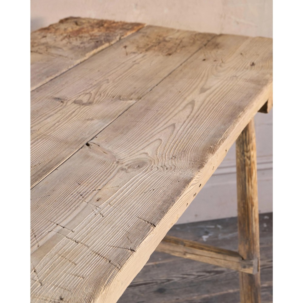 A primitive rustic dining table constructed from weathered pale timber with a rough-hewn plank top and diagonal cross-braced legs. View XIV