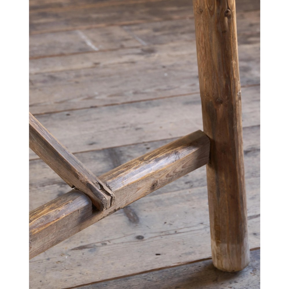 A primitive rustic dining table constructed from weathered pale timber with a rough-hewn plank top and diagonal cross-braced legs. View XV