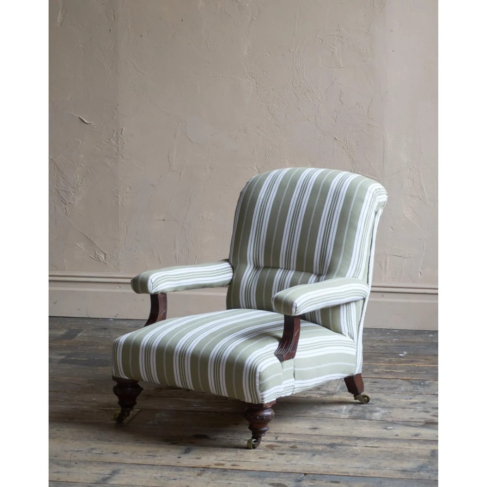 An oversized 19th-century open armchair upholstered in sage green and white striped fabric with carved mahogany arm supports and turned legs on brass castors.