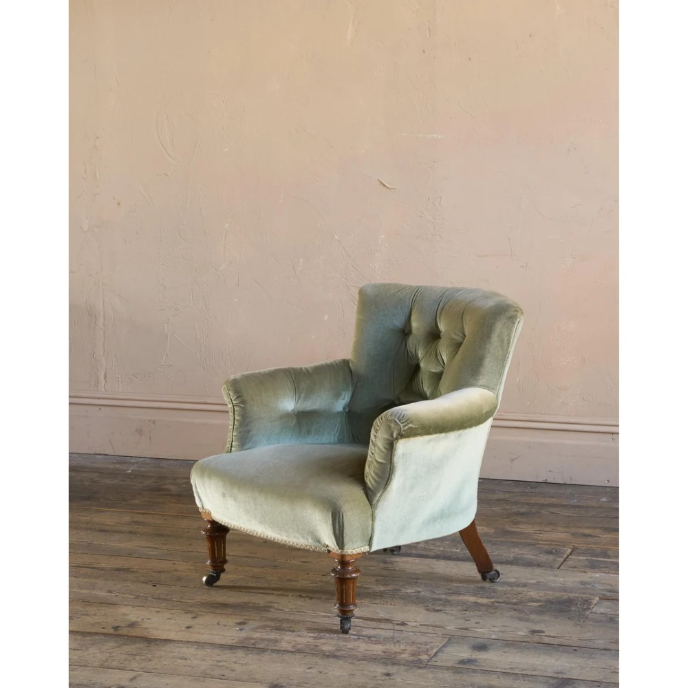 A 19th-century Victorian low armchair upholstered in light sage green velvet with a button-back design and turned wooden front legs on brass castors.
