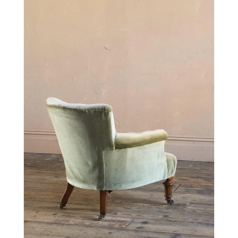 A 19th-century Victorian low armchair upholstered in light sage green velvet with a button-back design and turned wooden front legs on brass castors. View V