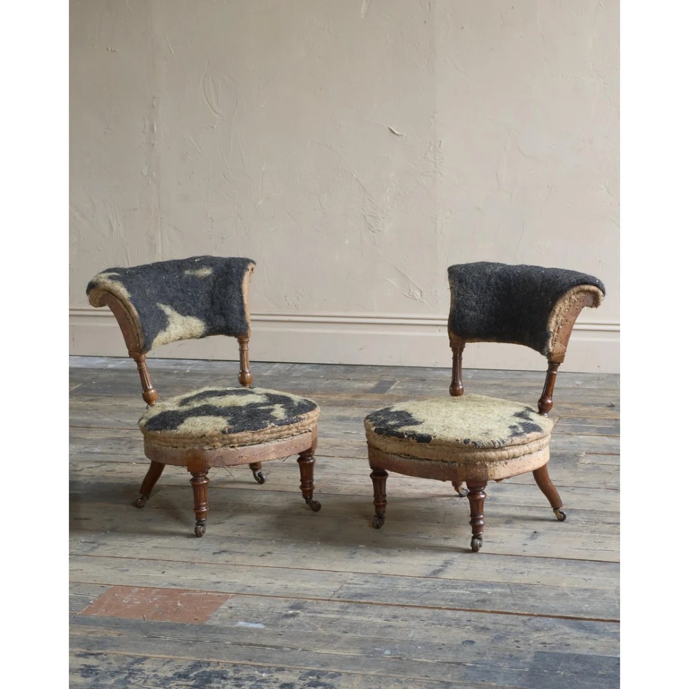 A pair of antique Victorian low-seated nursing chairs featuring rounded seat frames, turned front legs on brass castors, and exposed inner upholstery stuffing.