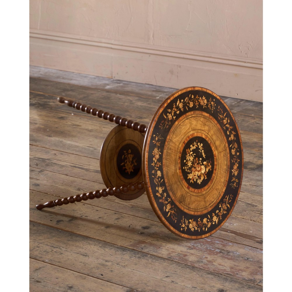 A circular warm brown wooden side table featuring floral marquetry inlays on the tabletop and lower shelf, supported by four bobbin-turned legs. View V