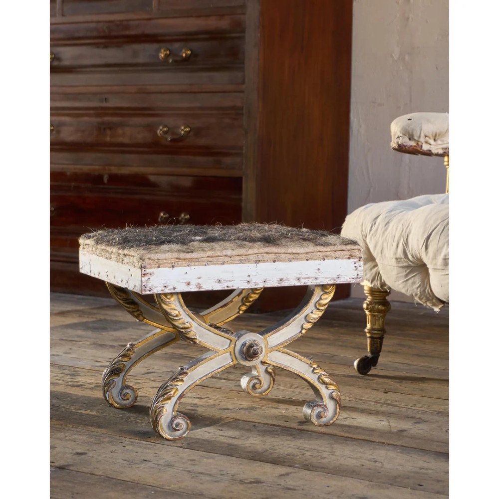 An antique curule X-frame footstool featuring carved scroll legs in grey paint and gilded finish with an upholstered top base.