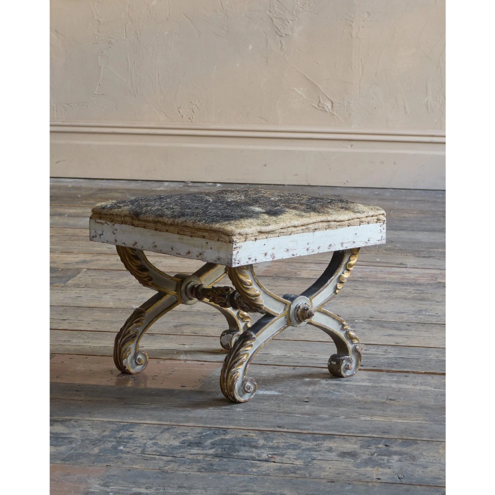 An antique curule X-frame footstool featuring carved scroll legs in grey paint and gilded finish with an upholstered top base. View V
