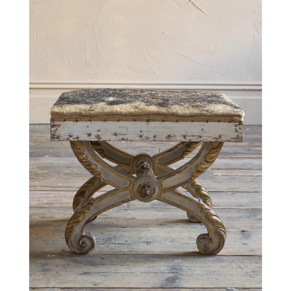 An antique curule X-frame footstool featuring carved scroll legs in grey paint and gilded finish with an upholstered top base. View VII