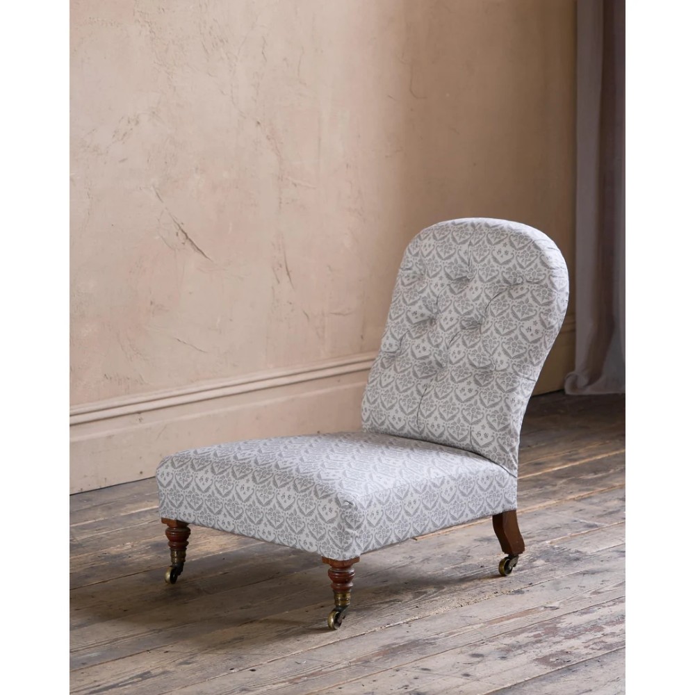 A low-seated Victorian armless slipper chair upholstered in grey patterned fabric with a button-back design and turned legs on brass castors.