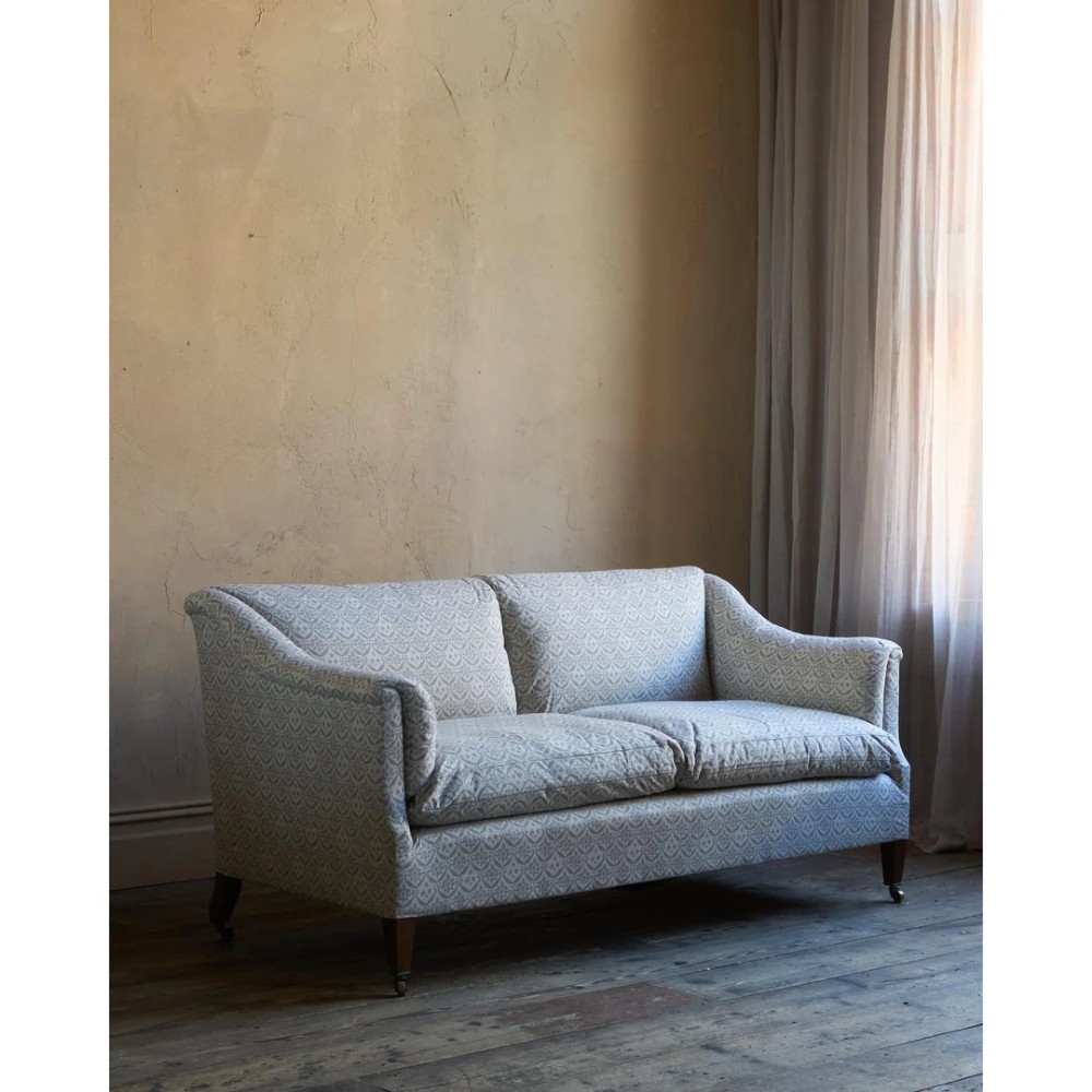 A two-seater Howard style sofa with sloped arms, grey geometric patterned upholstery, turned timber front legs, and brass castors.
