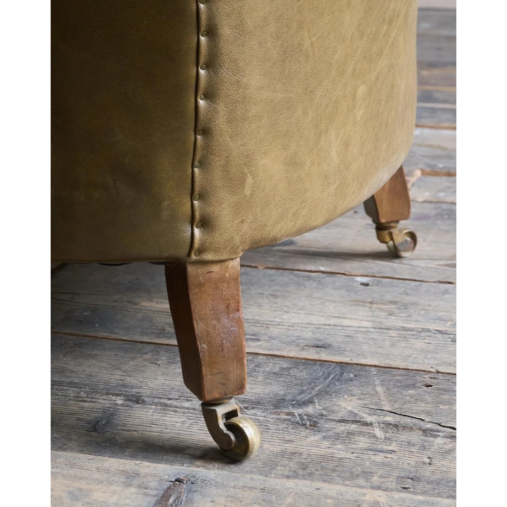 A deep-buttoned olive green leather armchair featuring turned wooden front legs with brass castors. View X