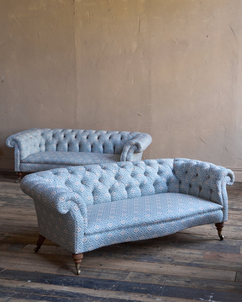 A pair of antique serpentine Chesterfield sofas featuring light blue patterned fabric, deep button-tufted backrests, rolled arms, and turned wooden legs on brass castors. View II