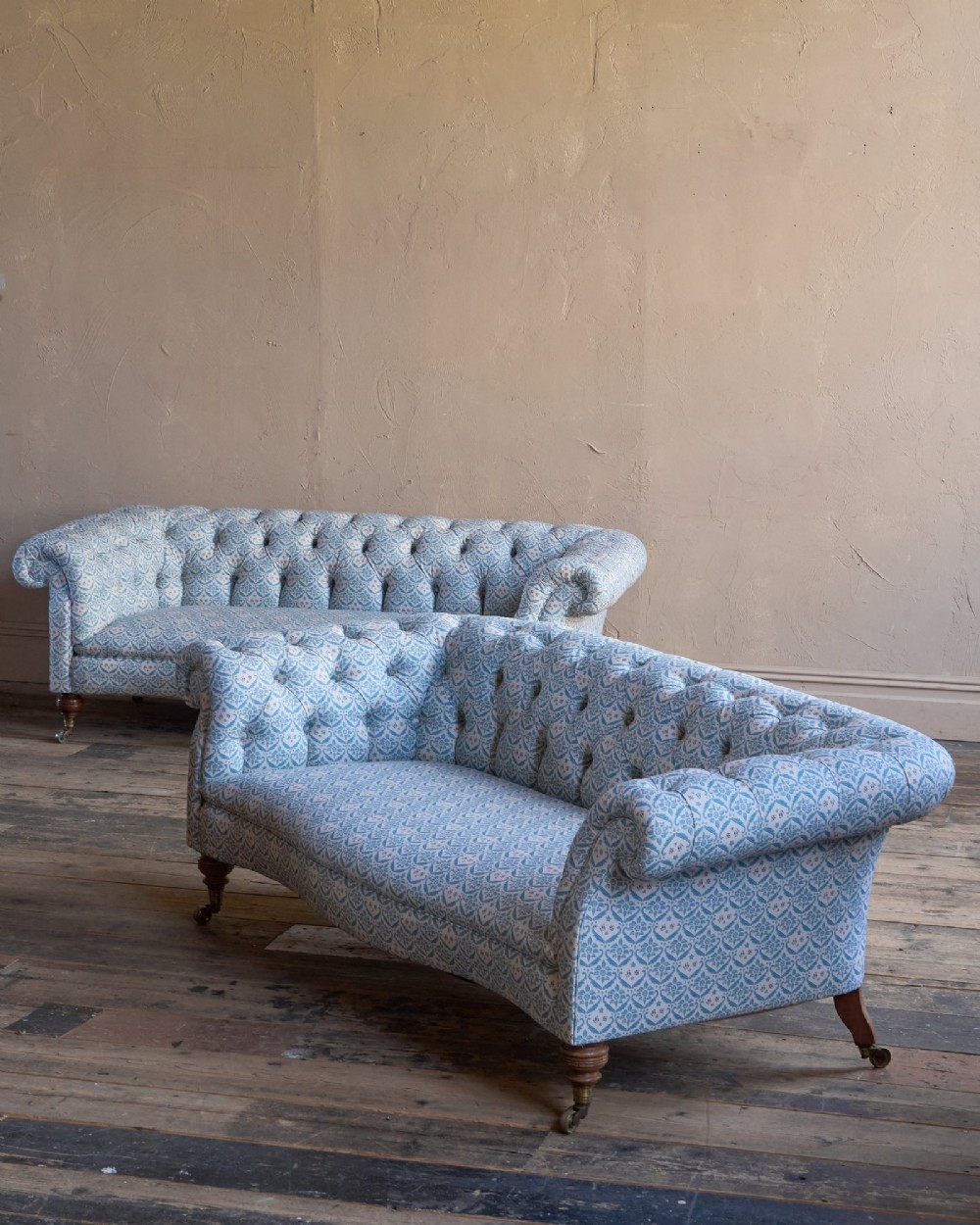 A pair of antique serpentine Chesterfield sofas featuring light blue patterned fabric, deep button-tufted backrests, rolled arms, and turned wooden legs on brass castors. View III
