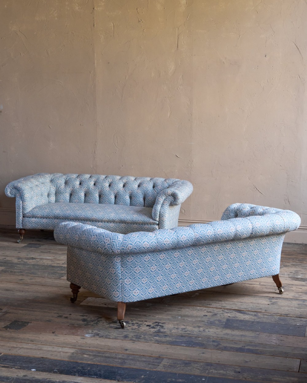 A pair of antique serpentine Chesterfield sofas featuring light blue patterned fabric, deep button-tufted backrests, rolled arms, and turned wooden legs on brass castors. View V