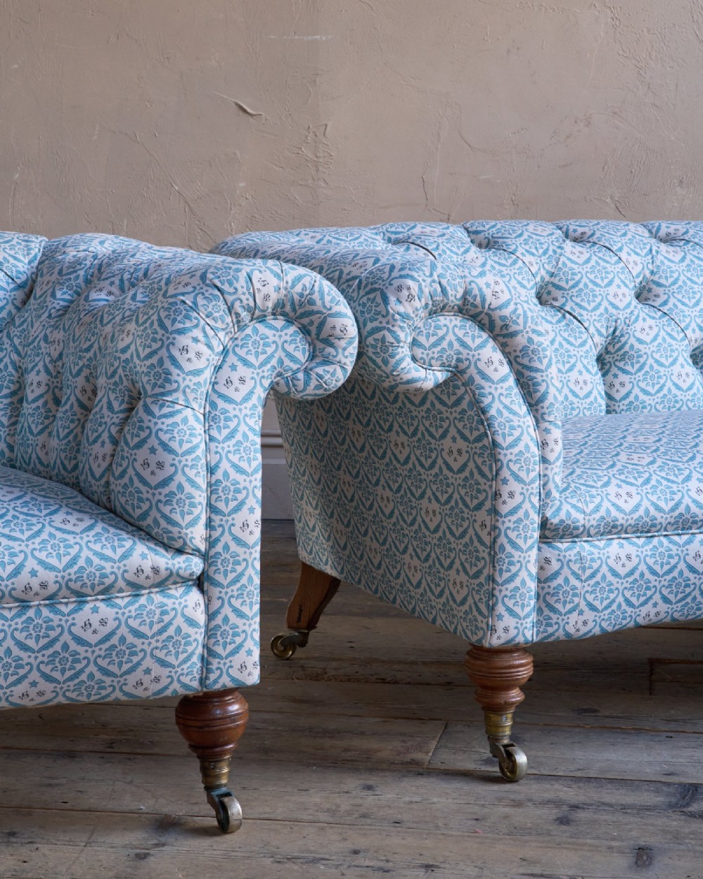 A pair of antique serpentine Chesterfield sofas featuring light blue patterned fabric, deep button-tufted backrests, rolled arms, and turned wooden legs on brass castors. View IX