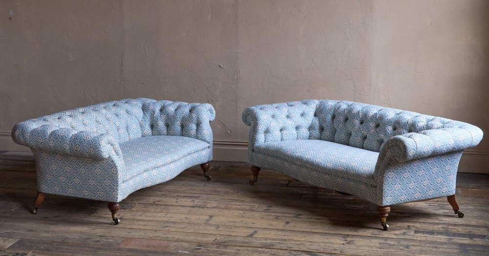A pair of antique serpentine Chesterfield sofas featuring light blue patterned fabric, deep button-tufted backrests, rolled arms, and turned wooden legs on brass castors. View X