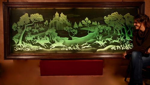 fig 1. A large rectangular dark wood frame housing an illuminated green sandblasted glass panel featuring a detailed hunting scene with horseback archers, hounds, stags, and a castle.