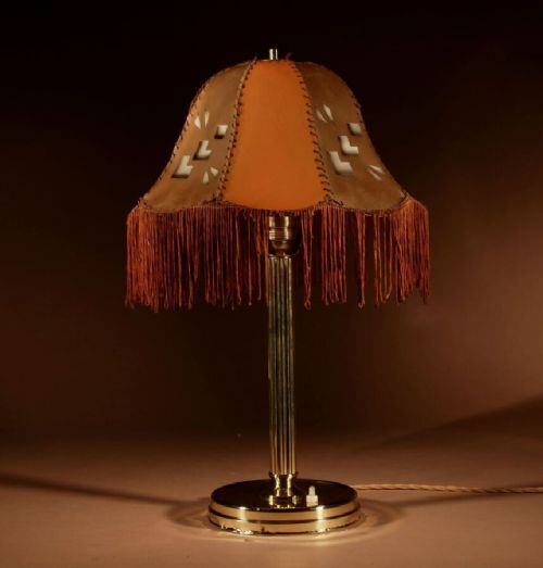 fig 1. An Art Deco brass table lamp featuring a reeded column, stepped circular base, and a tan leather shade with whipstitching, geometric cut-outs, and long amber fringe.