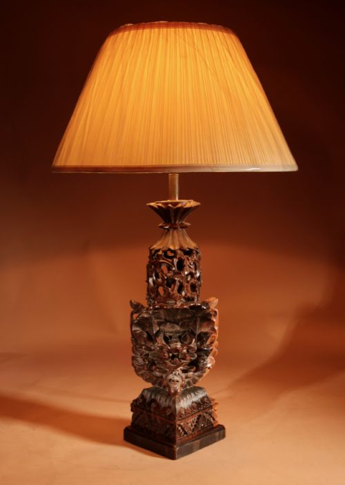 fig 1. A carved dark hardwood table lamp featuring a detailed mythological mask motif and pierced lattice section, topped with a pleated beige fabric shade.