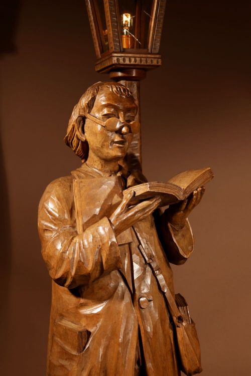 fig 1. A tall carved wooden sculpture depicting a scholar reading a book under a streetlamp mounted atop a miniature bookshelf base.