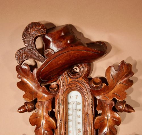 fig 1. A carved walnut wall-hanging aneroid barometer with a thermometer scale, featuring oak leaves, acorns, crossed rifles, and a carved hat top motif.